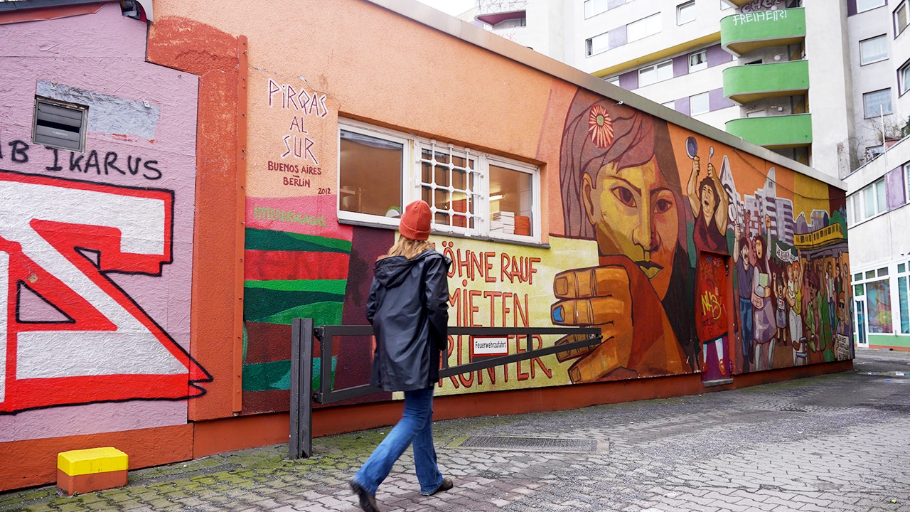 Eine ehemalige Stipendiatin der Rosa-Luxemburg-Stiftung läuft durch den Kiez in dem sie sich politisch engagiert. An der Wand ist ein großes Graffiti zu sehen, das zu hohe Mieten anprangert.  