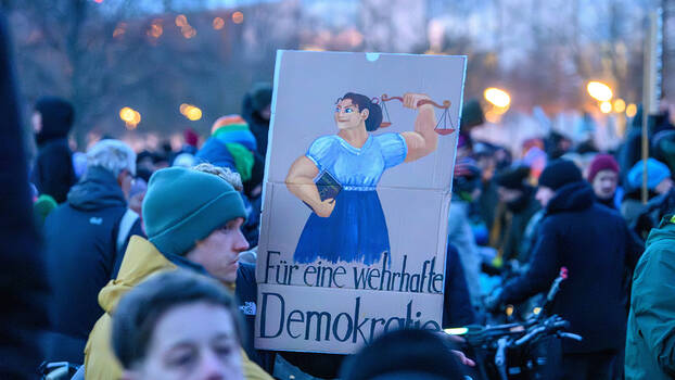 Demonstranten halten ein Schild, darauf eine Justizia-Figur mit einem Grundgesetz unter dem Arm und einem Schriftzug "Für eine wehrhafte Demokratie"