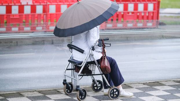 Seniorin sitzt mit Regenschirm auf einem Rollator auf der Straße